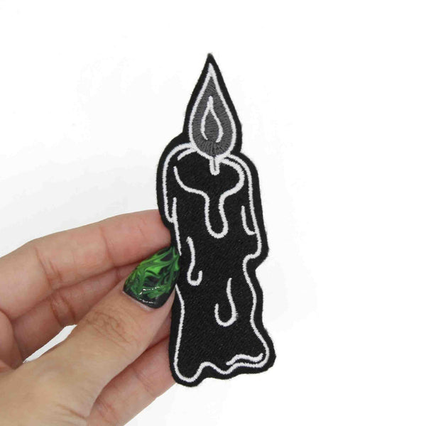 An embroidered patch of a lit melting black candle with white and grey details. Shown held for scale 
