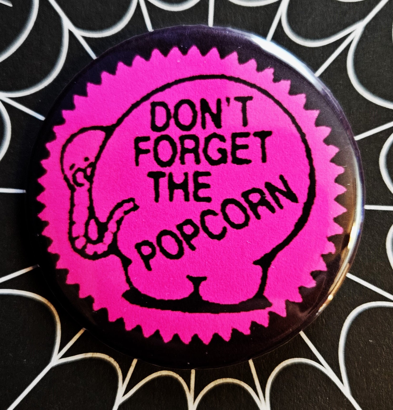 1.25” round button with image of pink zigzag border sticker with illustration of elephant with “Don’t forget the popcorn” message 