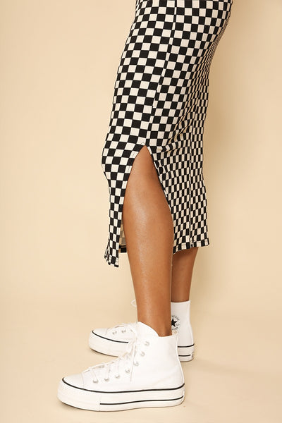 A model wearing a black and creamy white checkerboard pattern ribbed knit dress with long sleeves and a mock neck. Ends below the knee and has doubler slide slits. Shown from side in close up of side slit 