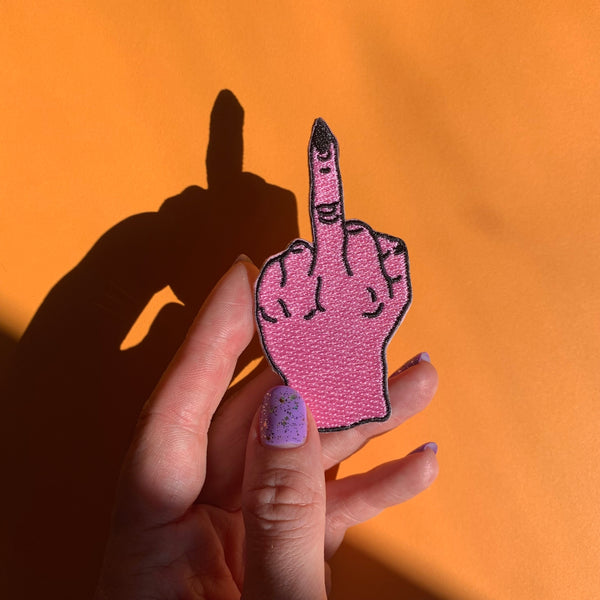 An embroidered patch of a bright pink hand giving the middle finger with pointy black fingernails and black detailing. Shown held for scale 