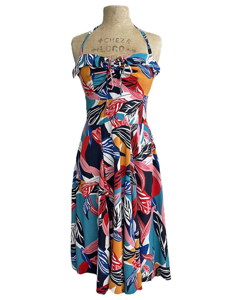 A halter dress in a bright blue, orange, pink, red, white, and black abstract tropical leaf pattern. It has a sweetheart neckline with exaggerated cuff details, cinched bodice, below the knee skirt, and adjustable halter tie neck. Shown on a dress form from the front