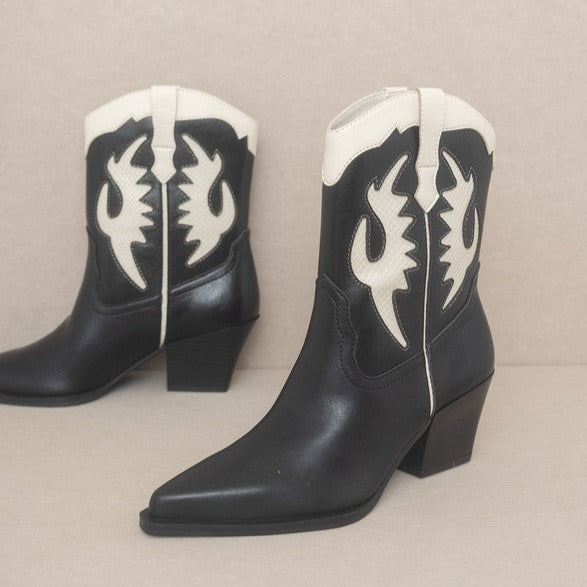 A pair of black pointed toe western style boots with pointed toes and 2” block heels. Black shiny faux leather with creamy white inset details. Shown from a three quarter angle