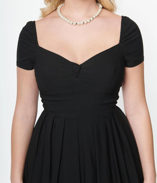 black stretch bengaline dress featuring short sleeves, an open sweetheart neckline, decorative button detail at the bust, fitted bodice, and pleated full just below the knee length skirt. showing torso close up on a model.