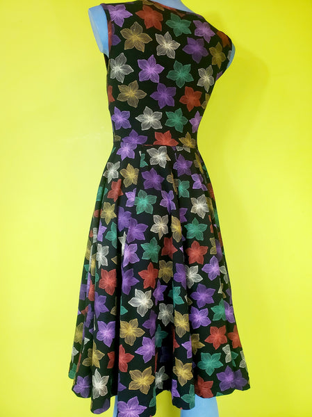 a mannequin wearing a fit and flare style dress in a cotton spandex knit with an all over pattern of fuchsia, purple, red, orange, creamy off-white, and teal blooming tropical flowers on a black background. Dress is faux-wrap style with a surplice bodice and v-neckline neckline. Full skirt ends below the knee and has side seam pockets. Seen from back