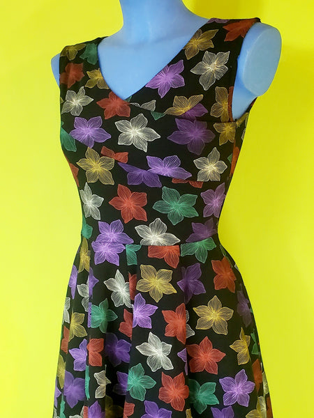 a mannequin wearing a fit and flare style dress in a cotton spandex knit with an all over pattern of fuchsia, purple, red, orange, creamy off-white, and teal blooming tropical flowers on a black background. Dress is faux-wrap style with a surplice bodice and v-neckline neckline. Full skirt ends below the knee and has side seam pockets. Seen from front