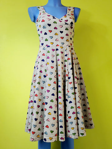 mannequin wearing cotton fit and flare sleeveless dress with surplice bodice and v neckline. Dress has a full knee length skirt and pockets. Warm light beige background with delicate vertical stripe pattern and all over staggered cartoony red, yellow, light blue, royal blue, purple, and pink heart pattern. Shown from the front