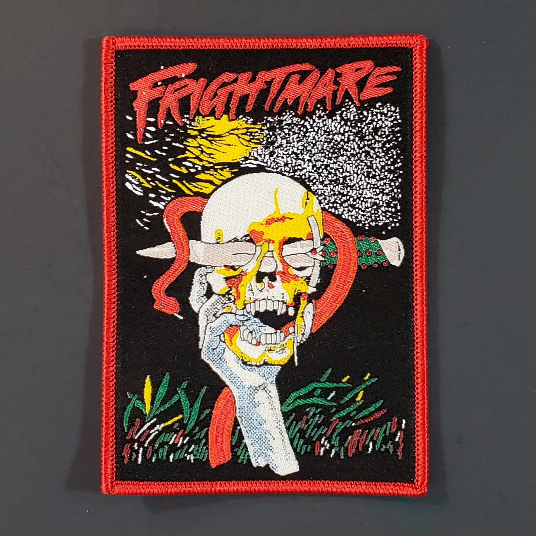 Rectangular red, yellow, green, grey, and black woven patch with illustration from the movie Frightmare depicting a hand holding a skull with a large blade stuck through the eye socket with a red snake coiled around it