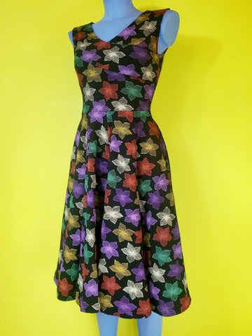 a mannequin wearing a fit and flare style dress in a cotton spandex knit with an all over pattern of fuchsia, purple, red, orange, creamy off-white, and teal blooming tropical flowers on a black background. Dress is faux-wrap style with a surplice bodice and v-neckline neckline. Full skirt ends below the knee and has side seam pockets. Seen from front