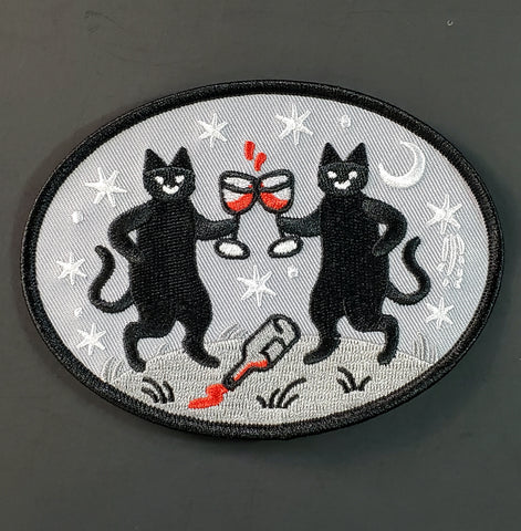 Oval twill embroidered patch of two black cats holding glasses of red wine with a spilled bottle at their feet surrounded by white moons and stars on a grey background