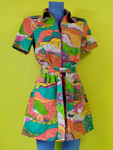 A mannequin wearing a stretch cotton mini dress with a rounded wide collar, short sleeves, and a bright purple plastic zipper running down the length of the dress. It has an intricate multicolored psychedelic pattern including pinwheels, apples, hands, birds, and other illustrations in pink, yellow, orange, blue, green, and white. Dress comes with a matching sash style belt. Shown from front