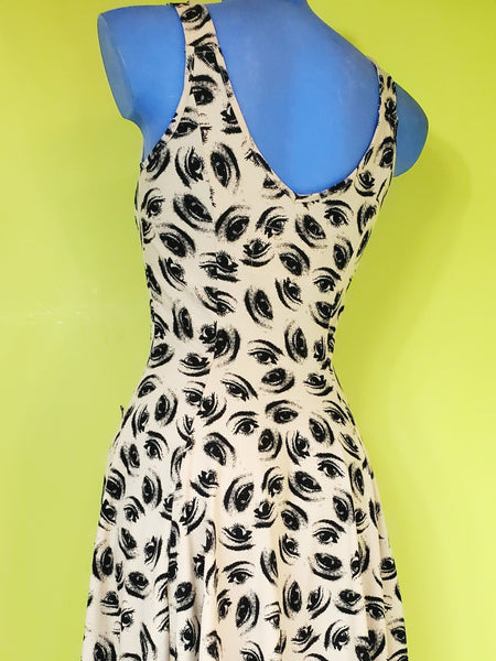 sleeveless stretch cotton knit dress in a black colored repeat pattern of hypnotic illustrated eyes against a warm cream background, featuring a v-neckline, fitted bodice with vertical seaming, scallop-top patch pockets, and a full flared knee length skirt. shown back view on a blue mannequin.