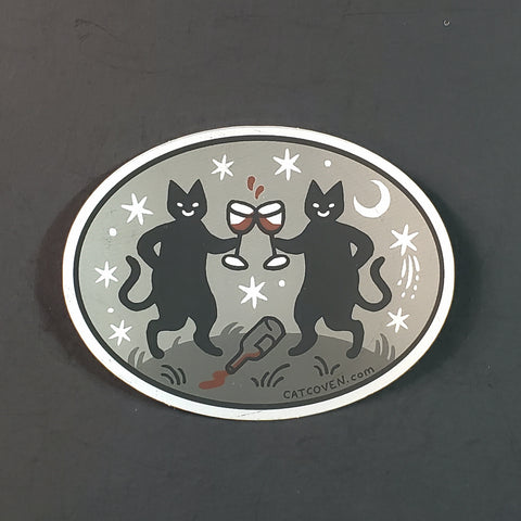 Oval vinyl sticker of two black cats holding glasses of red wine with a spilled bottle at their feet. On a metallic grey background with white moons and stars