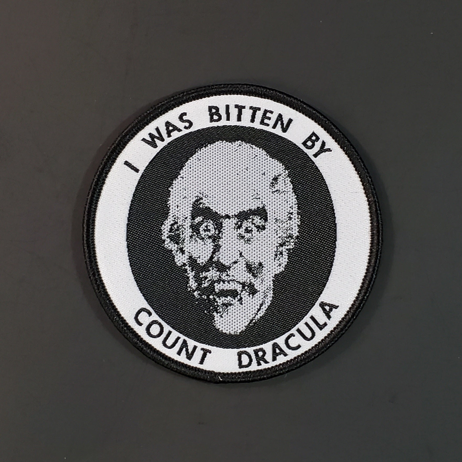 Woven black, white, and grey round patch with embroidered black border and illustration of vampire with “I WAS BITTEN BY COUNT DRACULA” written around it