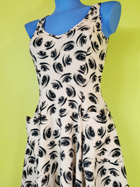 sleeveless stretch cotton knit dress in a black colored repeat pattern of hypnotic illustrated eyes against a warm cream background, featuring a v-neckline, fitted bodice with vertical seaming, scallop-top patch pockets, and a full flared knee length skirt. shown on a blue mannequin.