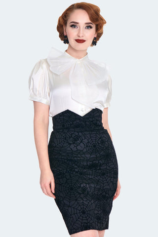 A model wearing a black-on-black flocked stretch taffeta pencil skirt with a rose and spiderweb pattern. It has a a v-shaped cinched cummerbund style waistband with matching diagonal button detail and a slit at the hemline. Shown from the front