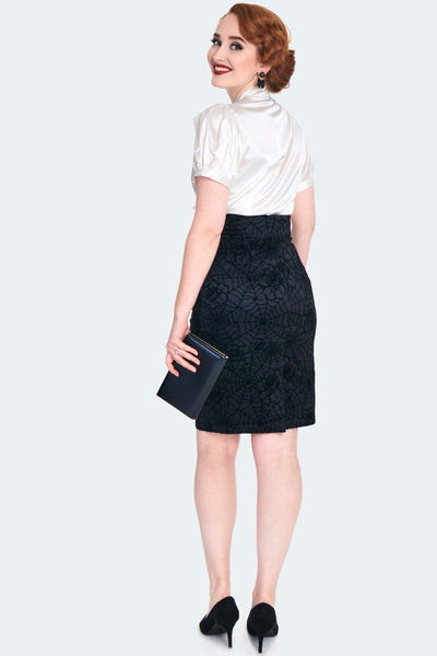 A model wearing a black-on-black flocked stretch taffeta pencil skirt with a rose and spiderweb pattern. It has a a v-shaped cinched cummerbund style waistband with matching diagonal button detail and a slit at the hemline. Shown from behind