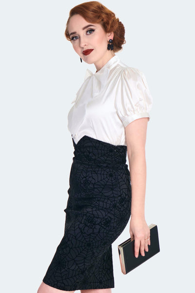 A model wearing a black-on-black flocked stretch taffeta pencil skirt with a rose and spiderweb pattern. It has a a v-shaped cinched cummerbund style waistband with matching diagonal button detail and a slit at the hemline. Shown from a three quarter angle 