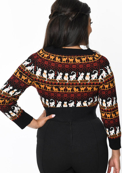 A model wearing a jacquard knit Fair Isle style design sweater with a scoop neck and three quarter sleeves. It has black ribbing at the neckline, cuffs, and bottom band. Pattern of red snowflakes and orange and white kitties with orange scarves and red collars against a black background. Shown from the back