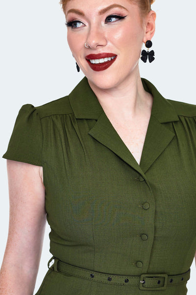 A model wearing a dark olive green shirtwaist dress with a v-neck and notched collar. It has gathering at the shoulders and slightly puffed cap sleeves. Hitting at the natural waist with a self belt and ends just above the knee with a slight flare. Seen from the front in close up of bodice