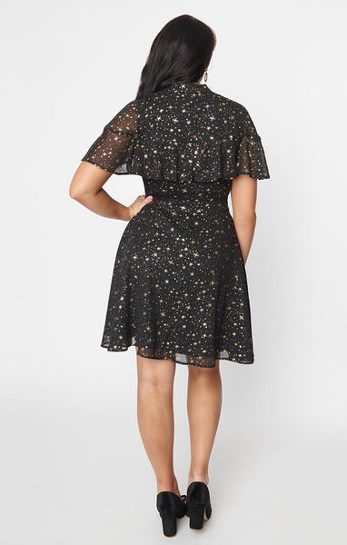 A black crepe chiffon dress with an all-over pattern of small gold stars and starbursts. It has a mandarin collar with keyhole and an attached caplet with a ruffled hem. The full skirt ends just above the knee. Shown from the back worn by a model