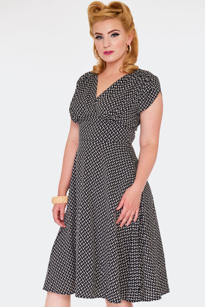 A model wearing a just below the knee length crepe fabric dress with a surplice style v-neckline, fitted waist, and tulip cap sleeves. Pattern of black and white zig-zag stripes with overlapping lines similar to basketweave. Shown from the front in a three quarter angle