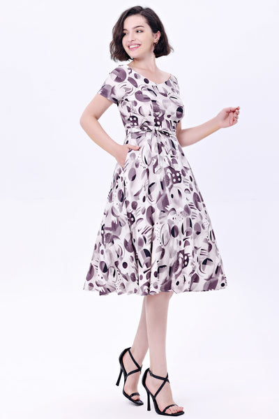 Model wearing a cocktail length short sleeved dress with a full gathered skirt and surplice style v neckline. It has a white background patterned with achromatic grey and black abstract shapes. Shown from three quarter angle