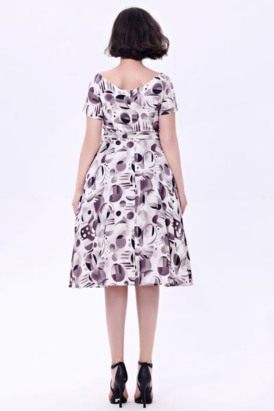 Model wearing a cocktail length short sleeved dress with a full gathered skirt and surplice style v neckline. It has a white background patterned with achromatic grey and black abstract shapes. Shown from back