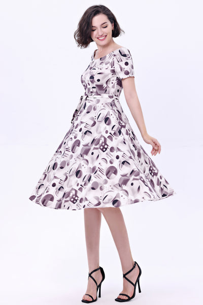 Model wearing a cocktail length short sleeved dress with a full gathered skirt and surplice style v neckline. It has a white background patterned with achromatic grey and black abstract shapes. Shown from three quarter angle with skirt in motion