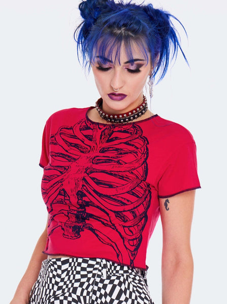Model wearing a cropped red t-shirt with a black screen printed image of a skeleton’s ribs on the front. Shirt has raw edge with black thread serged edges. Seen from three quarter angle