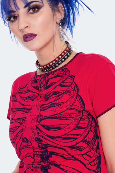 Model wearing a cropped red t-shirt with a black screen printed image of a skeleton’s ribs on the front. Shirt has raw edge with black thread serged edges. Seen from front in close up