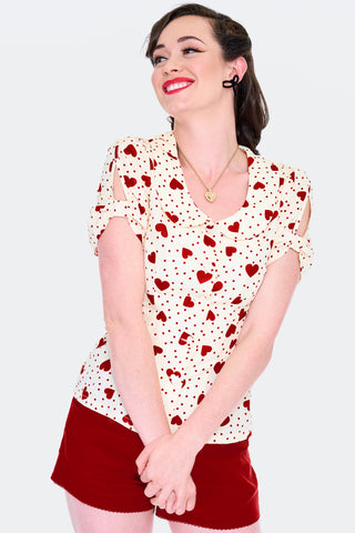 Model wearing short sleeved cream colored blouse with red dot and heart pattern. Has scooped Peter Pan collar, peekaboo detail at sleeves, and heart shaped fabric covered buttons. Shown from front