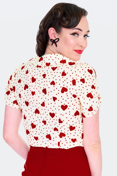 Model wearing short sleeved cream colored blouse with red dot and heart pattern. Has scooped Peter Pan collar, peekaboo detail at sleeves, and heart shaped fabric covered buttons. Shown from back