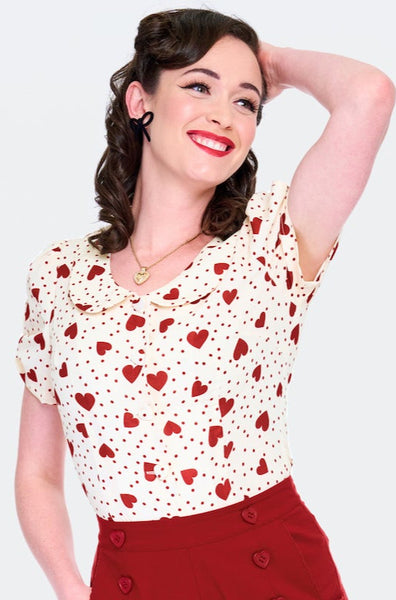 Model wearing short sleeved cream colored blouse with red dot and heart pattern. Has scooped Peter Pan collar, peekaboo detail at sleeves, and heart shaped fabric covered buttons. Shown from front with arm raised 
