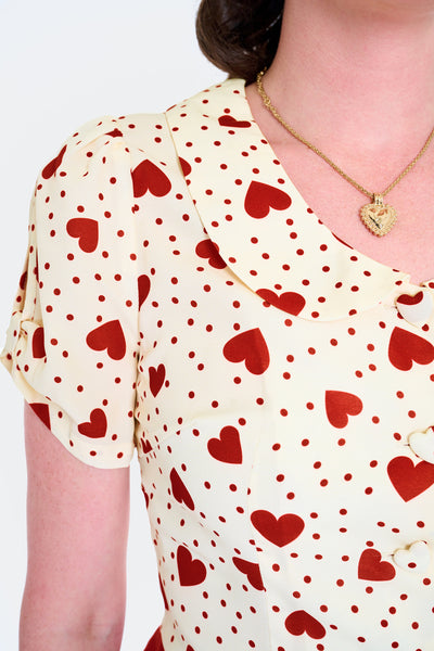 Model wearing short sleeved cream colored blouse with red dot and heart pattern. Has scooped Peter Pan collar, peekaboo detail at sleeves, and heart shaped fabric covered buttons. Shown from front in close up