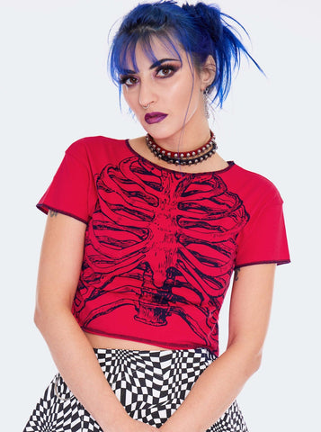 Model wearing a cropped red t-shirt with a black screen printed image of a skeleton’s ribs on the front. Shirt has raw edge with black thread serged edges. Seen from front