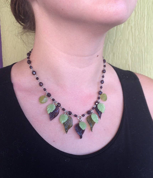 Faceted amethyst glass bead and silver toned metal link necklace with dangling amethyst, and pale and bottle green glass leaves, shown worn by model