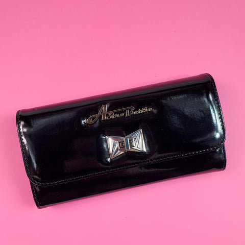 A rectangular tri-fold wallet with a shiny black vinyl exterior. It t has a large silver Astro Bettie logo on its exterior with a silver metal clasp in an Art Deco style