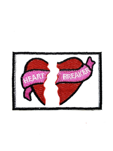 white canvas patch picturing a red heart broken in two with a hot pink banner running between each side reading “HEART BREAKER” with black stitched border