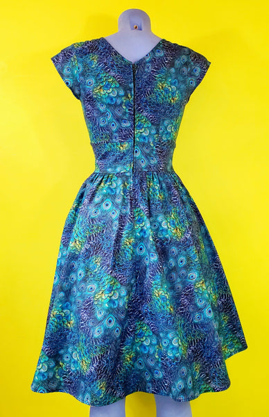 cap sleeve cotton dress in a green and blue peacock feather print with a surplus gathered neckline, wide gathered waist, and a just past the knee full skirt, shown back view on a mannequin
