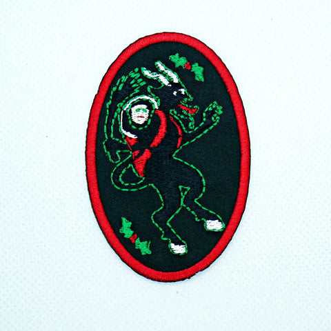 An oval black twill patch with a red border with Krampus embroidered in green and red and pictured with a misbehaving child over his shoulder