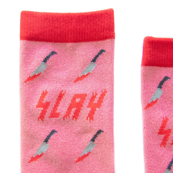 A pair of socks pink crew socks in an all-over bloody kitchen knife pattern with the word “SLAY” in a red bloody font shown in close up