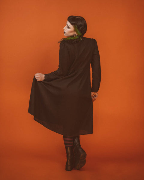A model wearing a long sleeved tea length linen shirtwaist dress with slightly puffed shoulders and princess seaming on the bodice. It has small matte black buttons down the front. The full skirt is slightly gathered. The model is shown from behind and is holding the full skirt up