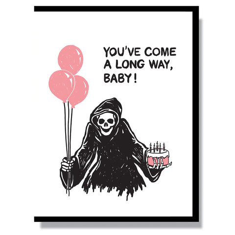 A rectangular greeting card with an image of the grim reaper holding a pink birthday cake and set of balloons with the message, “you’ve come a long way baby!”