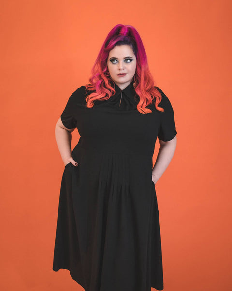 A model wearing a black 40s style short sleeved midi dress. elongated platter style collar and a narrow keyhole opening at the sternum with gathered detail and hook and eye closure. There is gathered pleating at the front of the full skirt. The model is plus size and has their hands in both side seam pockets
