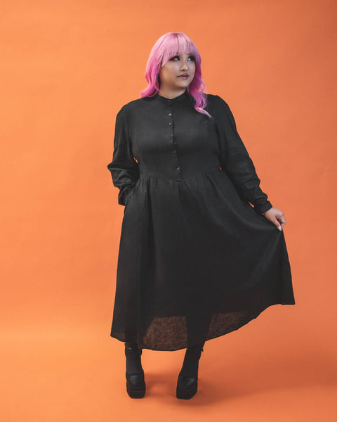 A plus size model wearing a long sleeved tea length linen shirtwaist dress with slightly puffed shoulders and princess seaming on the bodice. It has small matte black buttons down the front. The full skirt is slightly gathered. The model has their hand in one pocket and is holding the full skirt out with the other hand
