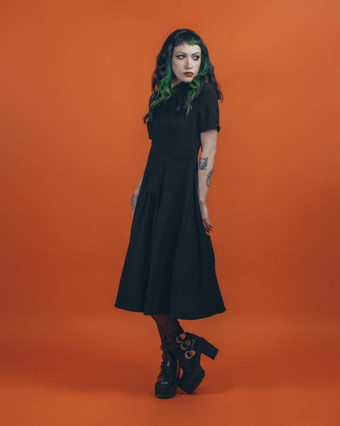 A model wearing a black 40s style short sleeved midi dress. elongated platter style collar and a narrow keyhole opening at the sternum with gathered detail and hook and eye closure. There is gathered pleating at the front of the full skirt. She is posing with her arms down 