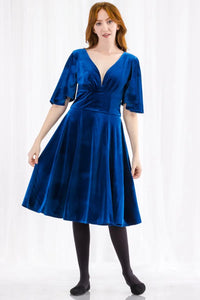 A royal blue velvet fit and flare dress with a deep plunging v sweetheart neckline, a wide banded waist, and elbow length flutter sleeves. Its skirt ends just past the knee. Shown on a model 