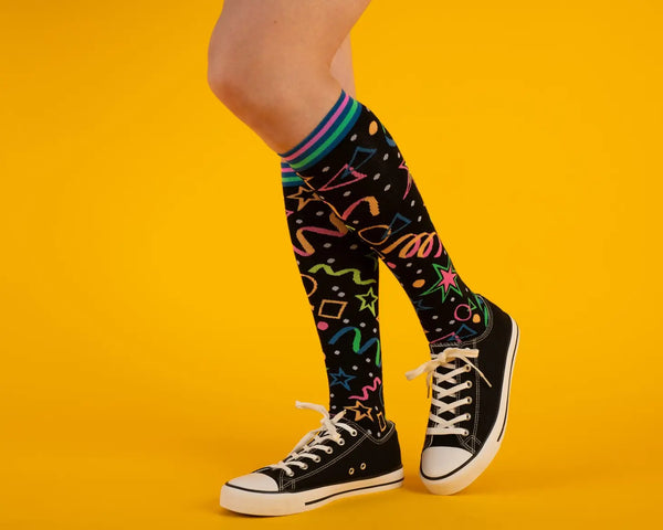 A pair of black knee socks with neon blue, green, and pink stripes at the cuff and blue toes & heels. There is a neon colored geometric pattern along the calf of each sock similar to an 80s bowling alley carpet. Shown on a model wearing black sneakers