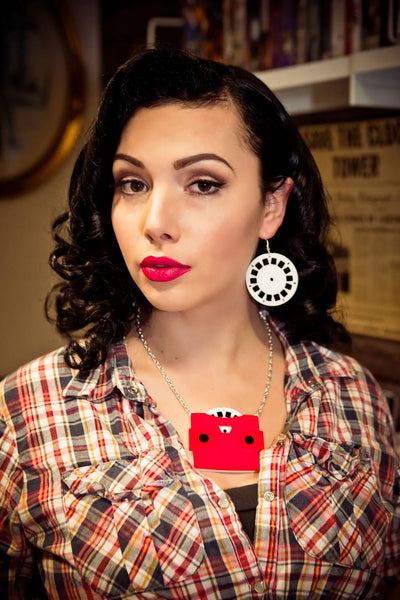A layered acrylic necklace in the shape of a view finder toy with a red body and black eye holes. It has a white and black view finder reel poking out of its top. On a silver plated curb link chain. Shown on a model wearing matching view finder reel dangle earrings 