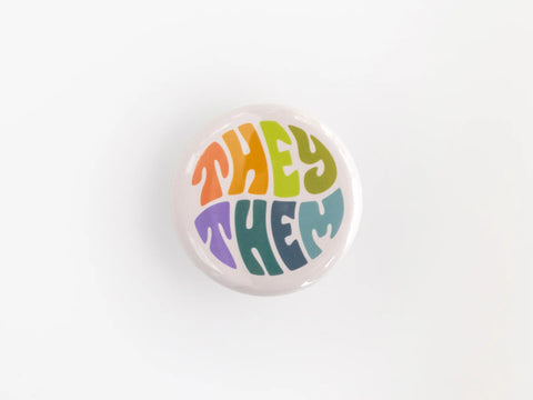 A 1.25” button with the pronouns “they” and “them”  in multicolored lettering